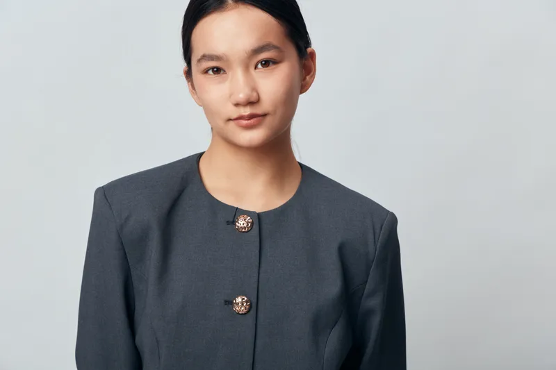 Young Asian woman in formal suit standing against light background looking at camera with calm expression for professional business portrait and confident career image.