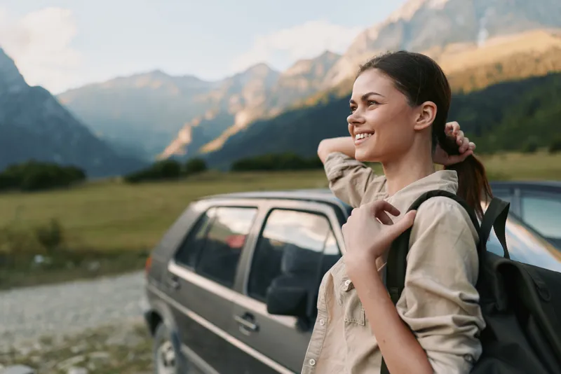 A smiling woman in casual clothes stands near a car in a mountain landscape, enjoying the outdoors and fresh air. Concept for lifestyle, travel, and freedom.