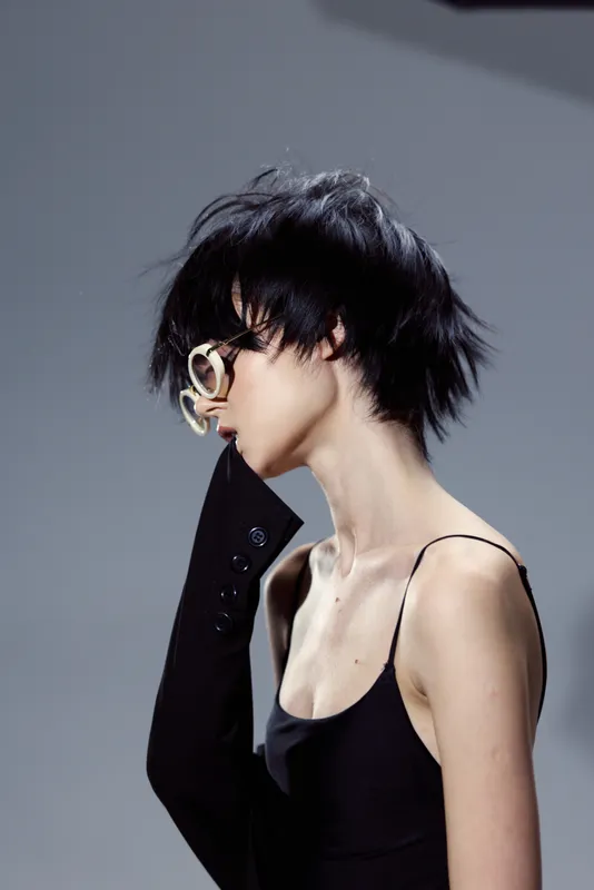 Profile of a thoughtful woman with short black hair wearing glasses and a black outfit in a minimalist studio setting
