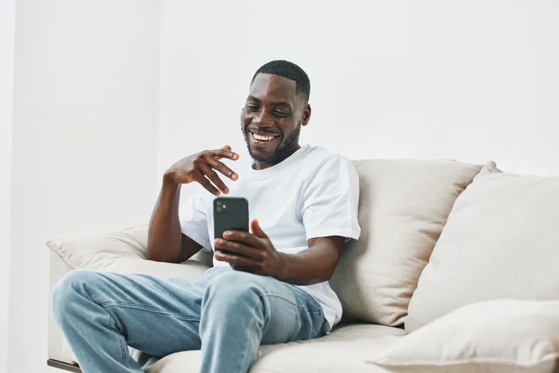 Happy young man enjoying his smartphone on a cozy couch, dressed casually in a white t shirt and blue jeans, creating a relaxed and joyful atmosphere
