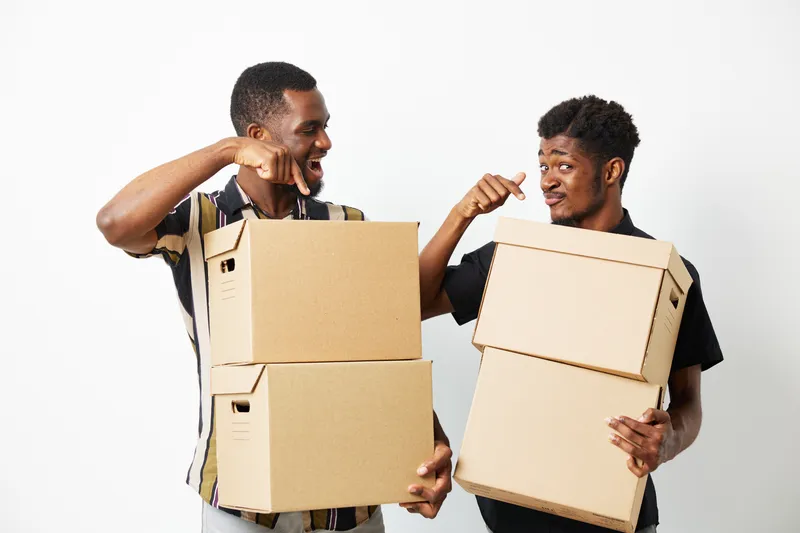 Two African American men joyfully interacting while holding cardboard boxes, showcasing teamwork and enthusiasm in a bright setting, perfect for deliveries or moving.