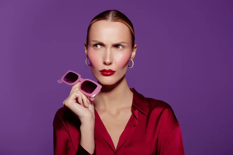 Confused woman in red blouse holds pink sunglasses near face on purple background. Studio portrait with expressive facial expression and stylish look. Concept of doubt, fashion, and attitude.