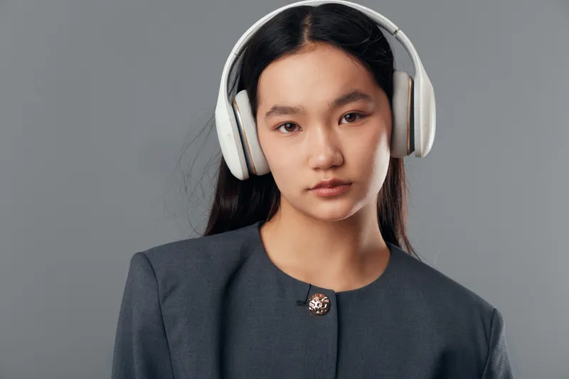 Young woman with long dark hair wearing white headphones and a gray blazer posing against plain gray background. Serious expression with natural makeup and clean minimal style.