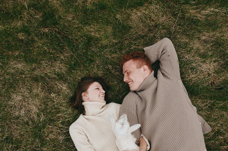 Casual couple outdoors on grass share warmth with a curious goat kid nearby, creating a cozy lifestyle moment. A candid scene captures affection, nature, and simple joy in a sunlit yard setting.