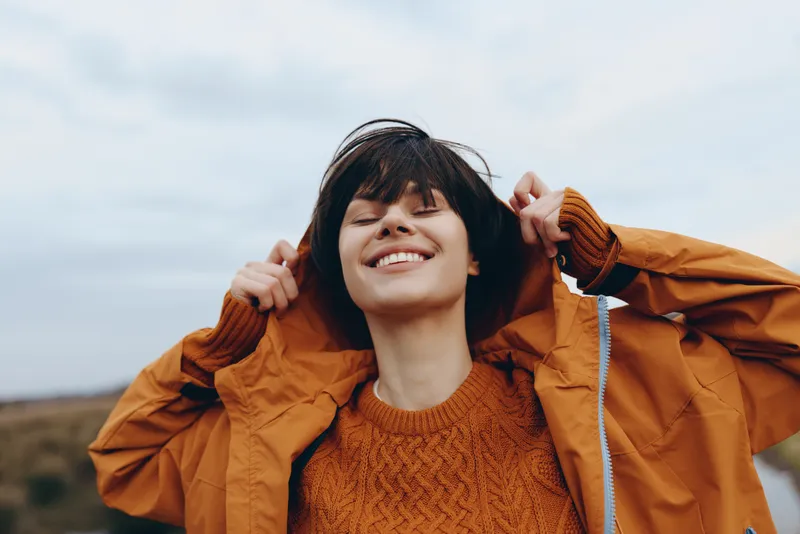 Woman outdoors lifestyle, jacket smile sweater cloudy wind casual portrait, orange jacket and knit sweater, hands adjusting collar, breezy daylight, relaxed pose, hills background