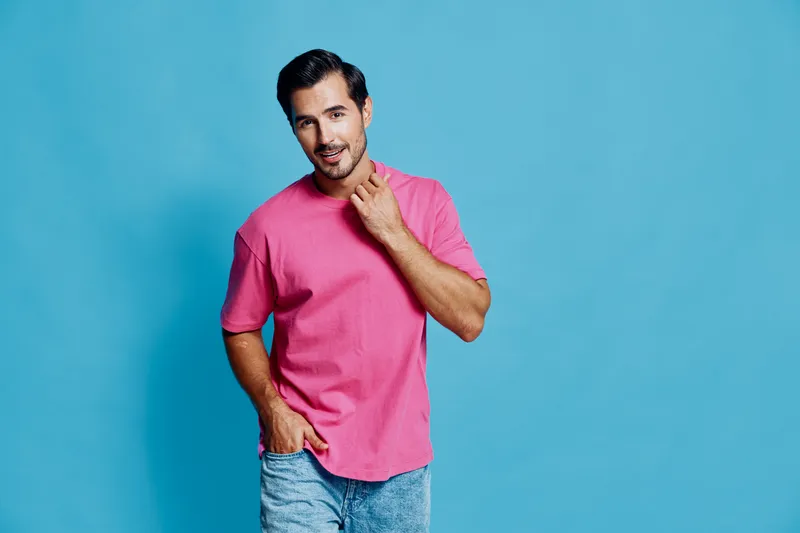 Full body smiling young man wearing pink t-shirt and denim shorts, posing against a bright blue background. Confident and cheerful lifestyle concept.