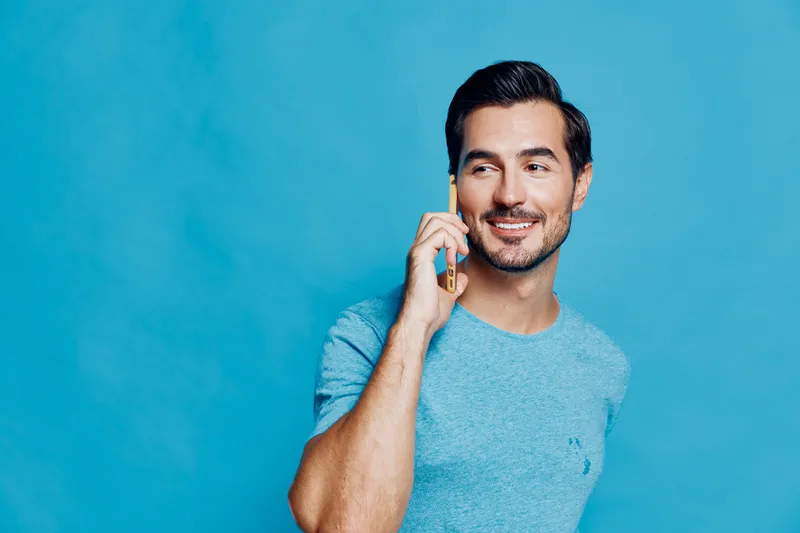 Charming young man smiling while talking on mobile phone, dressed in a casual blue t-shirt, standing against a vibrant blue background in a relaxed pose.
