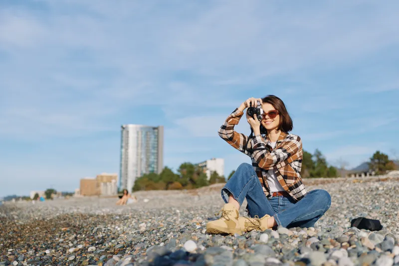 Woman in plaid shirt, jeans and boots sits on a pebbled beach, using a camera to capture the scene under a bright sky with distant urban skyline and calm sea.