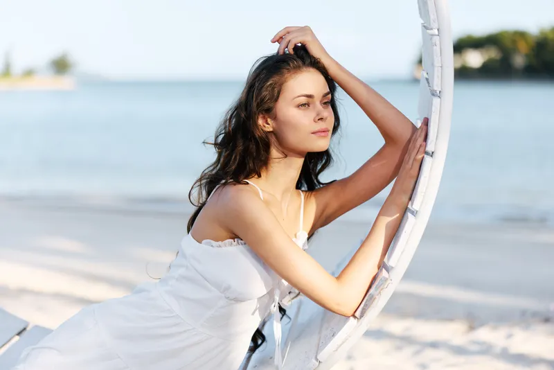 A woman in a white sundress leans against a weathered circular frame on a sunlit beach, gazing into the distance with calm poise as waves roll ashore.