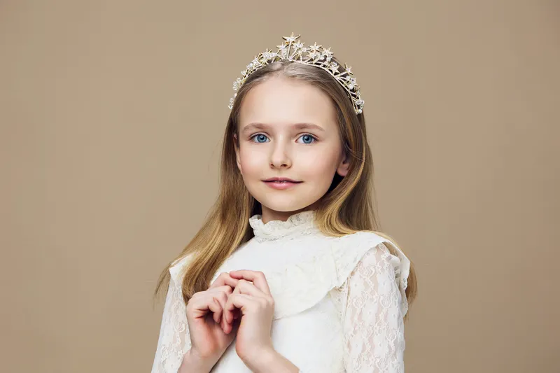 Charming portrait of a young girl in a white dress with a crown, showcasing innocence and grace against a soft beige background