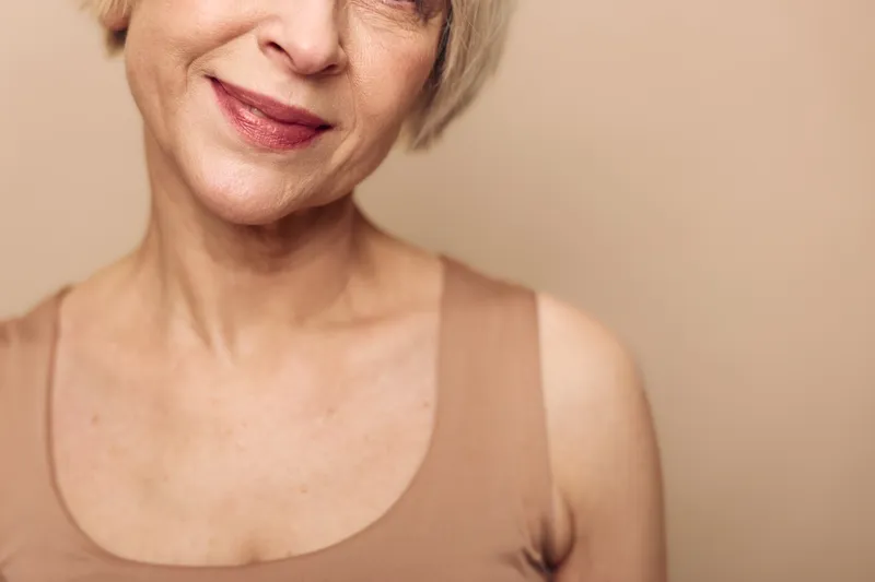 Close-up of smiling mature woman with short blonde hair wearing beige tank top against neutral background. Concept of natural beauty, aging gracefully, and confident lifestyle.
