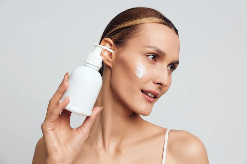 Woman holds skincare serum bottle near her facial area applying cream for beauty routine treatment, fresh skin hygiene care, calm expression, minimal studio shot on gray background