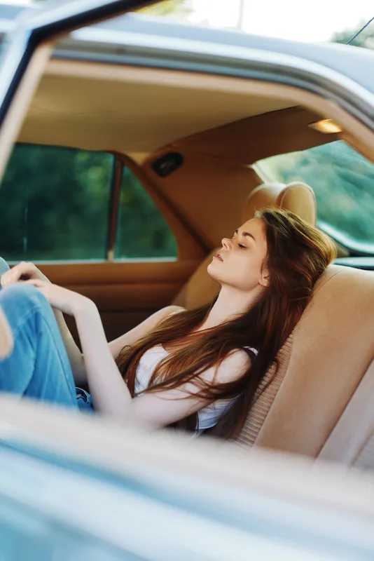 Young woman relaxing in a vintage car, wearing casual jeans and a white top, expressing calmness against a natural green background.