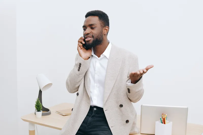 Smiling man with dark skin talking on mobile phone in modern office, dressed in beige blazer and white shirt, stand near desk with lamp and plant, casual business style, workspace environment, lifesty