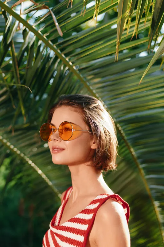 A stylish woman wears round sunglasses and a striped top, standing among lush palm fronds under bright sunlight, exuding a relaxed tropical vibe and carefree vacation mood.