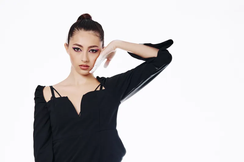 Young stylish woman with fashionable makeup posing confidently in elegant black dress. Clean white background highlights her beauty, fashion, and creative modern look. Studio photography.