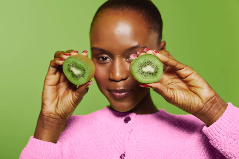 Young black woman with dark skin holds two halves of fresh kiwi fruit near face on green background. Concept of healthy lifestyle, natural food, and fresh fruit nutrition.