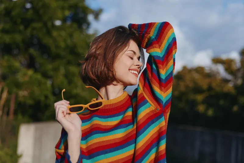 Young woman in mid twenties wearing a rainbow sweater and casual outfit, smiling with sunglasses in hand, short hair, outdoor portrait in sunlit park for lifestyle fashion.
