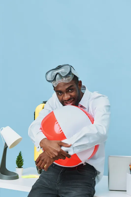 Playful young African American man in casual shirt with snorkeling mask holding a colorful beach ball, standing in a bright modern office with playful summer accessories, concept of fun, summer, vacat