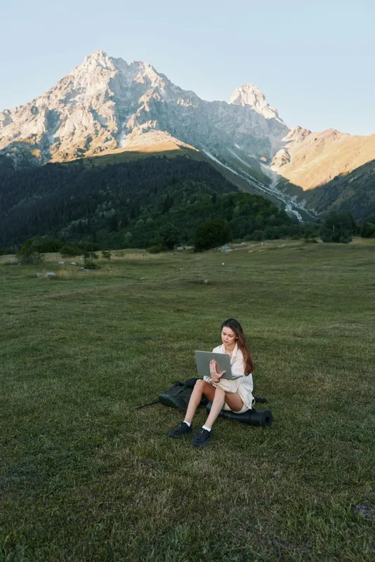 A focused student sits on a grassy field with a laptop, surrounded by rugged mountains. She studies outdoors, enjoying fresh air and sunlight, creating a calm and productive learning atmosphere.