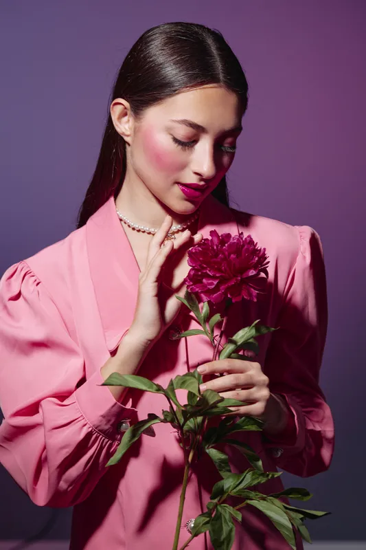 Young woman in a pink dress holding and smelling a dark red flower. She has glossy skin and makeup on a purple background. Creative beauty concept with elegant fashion style and soft lighting.