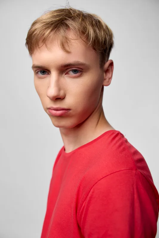 Young man in a red shirt gazes over his shoulder with a serious expression, studio lighting highlights cheekbones, clean background, fashion mood, contemporary casual photography style without