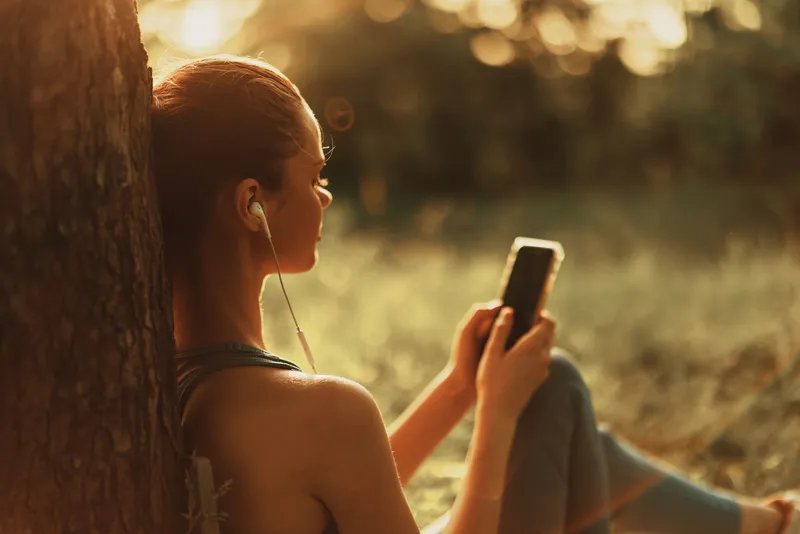 Relaxed young woman enjoying music outdoors in nature during sunset, wearing casual attire, embodying tranquility and leisure with earphones and smartphone