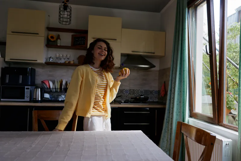 Cheerful young woman in a bright kitchen smiles as she holds an orange, wearing a yellow cardigan and striped shirt.