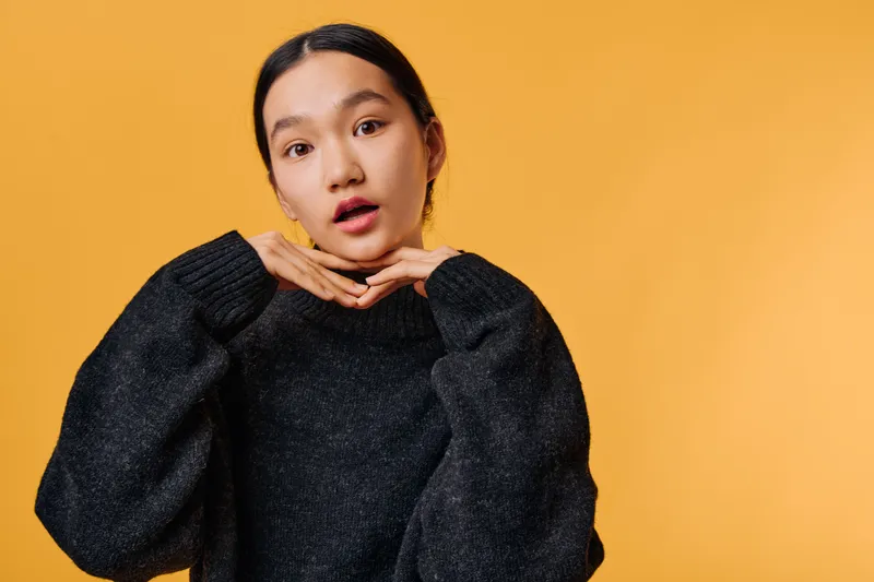 Young woman with dark hair in black sweater posing with hands under chin on orange background for portrait, emotion, and fashion style with confident expression indoors.