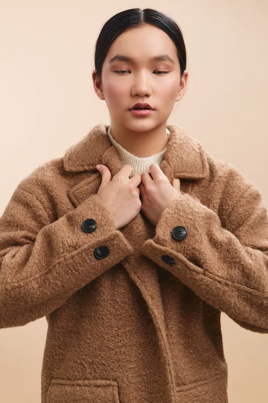 Asian woman adjusts her coat in a neutral studio setting, exuding calm confidence and poised style as she focuses on the task at hand and presents a minimalist, fashion oriented vibe.