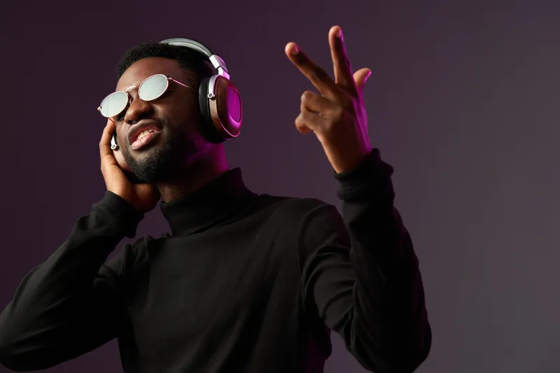 Stylish young Black man enjoying music with headphones and sunglasses in a modern dark background, expressing joy and confidence in a relaxed urban vibe