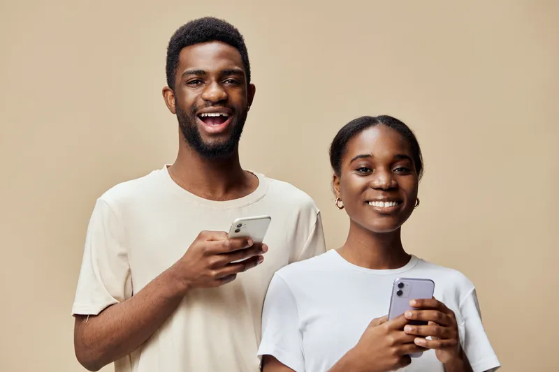 Happy young couple smiling while using smartphones, wearing casual outfits on a neutral background, representing connection and modern communication