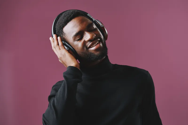 Young Black man enjoying music with headphones, wearing a stylish black turtleneck, smiling joyfully against a vibrant purple background