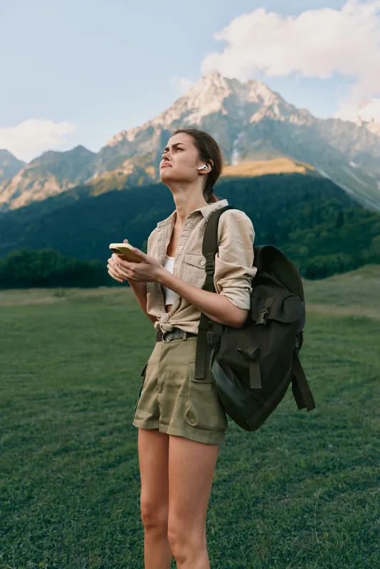 A young woman with a backpack stands in a green meadow, looking up at distant mountains while checking her phone, capturing a moment of exploration and solitude.