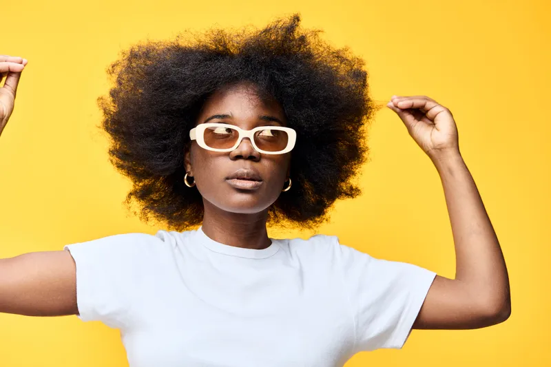 A young Black woman with natural textured hair, wearing stylish white sunglasses and a classic white t shirt, posing confidently against a vibrant yellow background
