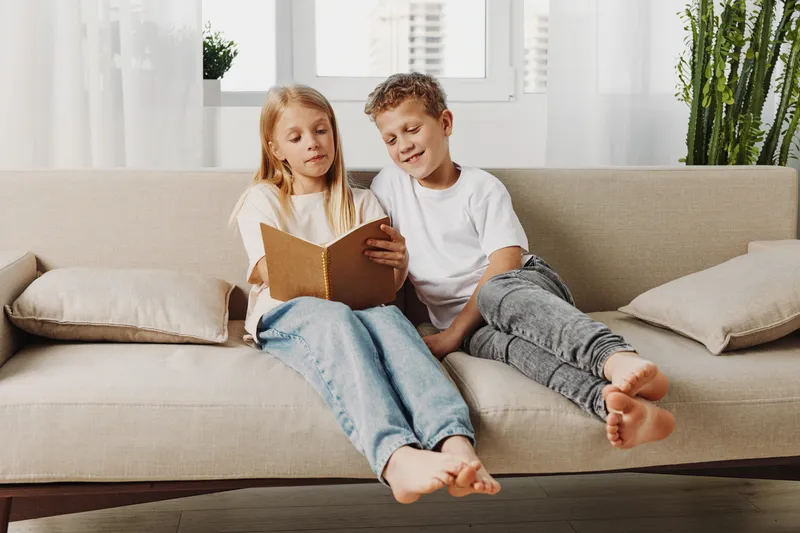 Two children sit barefoot on a beige sofa reading a book together in a bright living room. The casual atmosphere shows friendship, learning, and relaxed family time indoors.