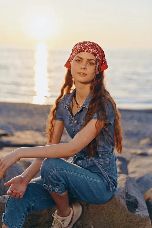 young woman with red headscarf sitting on rocks near sea during sunset in casual denim outfit, relaxed and calm mood outdoors