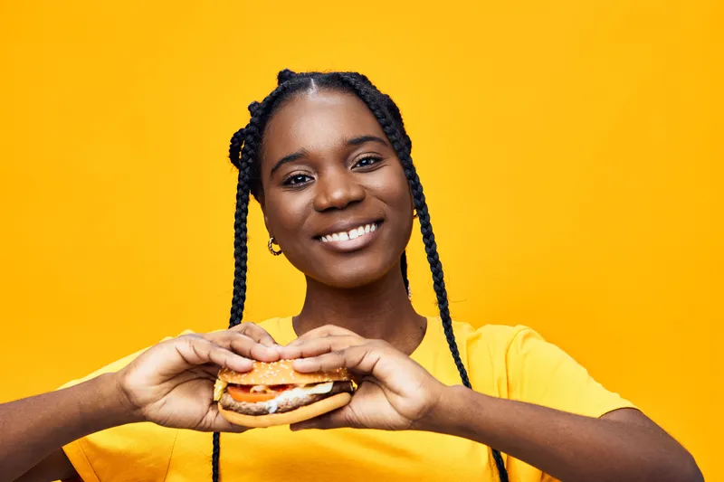 Happy young woman holding a delicious burger against a vibrant yellow background, showcasing healthy eating and enjoyment of comfort food