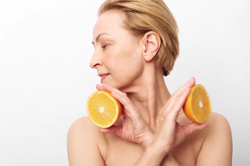 Mature woman with short blonde hair holds two orange halves near her face against white background. Concept of natural beauty, healthy lifestyle, skincare, and vitamin c benefits.