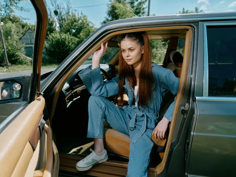 Stylish young woman in denim outfit sitting in a car, exuding confidence and joy in a sunny outdoor setting with greenery.