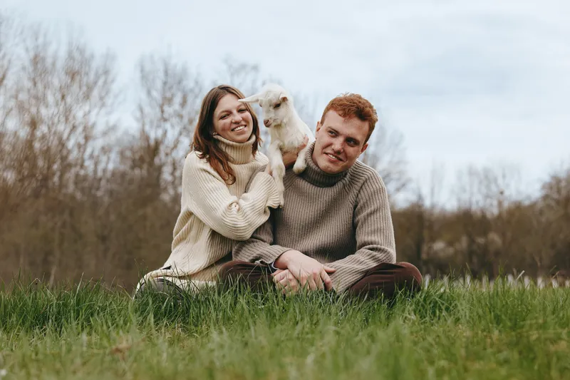 Smiling couple sitting on grass outdoors with a playful goat kid on a cloudy day. Rural lifestyle, bond, nature, and animal companionship. Soft natural light evokes warmth and relaxed, wholesome