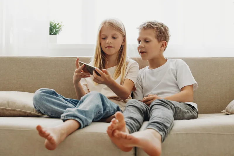 Two children sitting on a beige couch watching a smartphone together. Casual home environment with natural light and relaxed mood. Concept of technology use and family bonding.