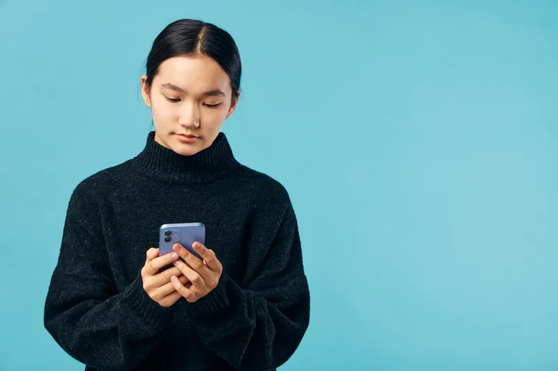 Teenage girl wearing black sweater using smartphone for texting or browsing with focused expression on blue background. Technology, communication and mobile device for social media and online chat.