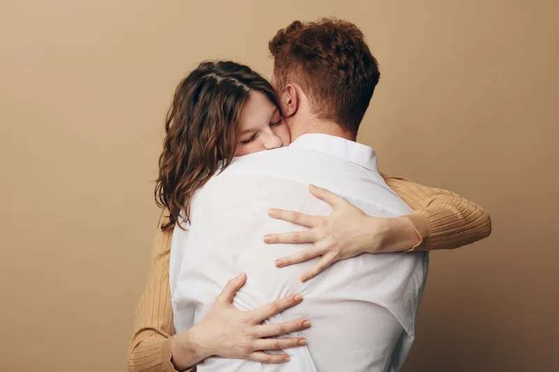 Young couple embraces tenderly with closed eyes, expressing love and comfort. Woman in brown sweater hugs man in white shirt against beige background. Concept of affection and intimacy.