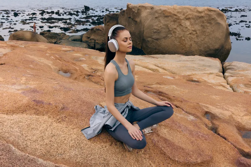 Young woman meditating on rocky beach, wearing sportswear, headphones, serene expression, with ocean background, promotes wellness and relaxation