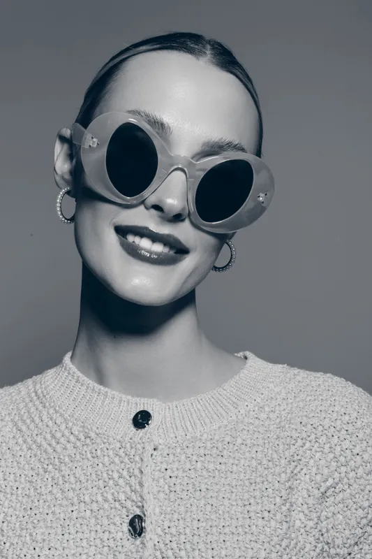 Smiling white woman with light skin wearing large round sunglasses and hoop earrings poses in a knitted sweater against grey background. Concept of fashion, style, and accessories.