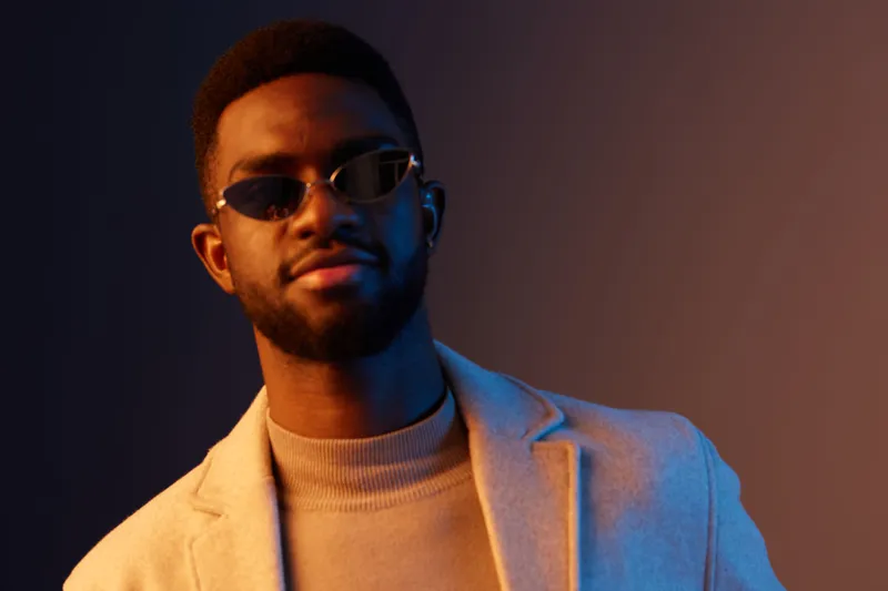 Stylish man wearing sunglasses in a beige coat against a gradient blue and orange background, exuding confidence and contemporary fashion