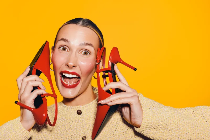 Happy woman with red high heels holds shoes near face, smiling widely on bright yellow background. Concept of fashion, style, and joyful emotions in studio lighting.