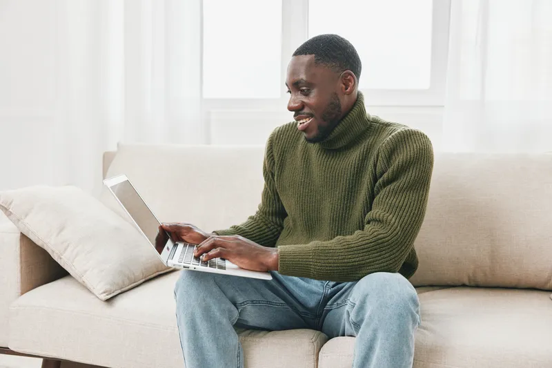 Smiling man in green sweater works on laptop at home, cozy atmosphere creating a sense of comfort, productivity, and relaxation in bright living space