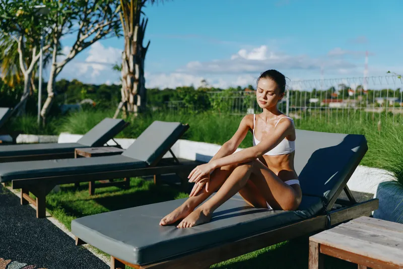 Young woman in white bikini sitting on a sun lounger outdoors, enjoying sunlight and fresh air. Concept of summer lifestyle, relaxation, and wellness in a natural setting.